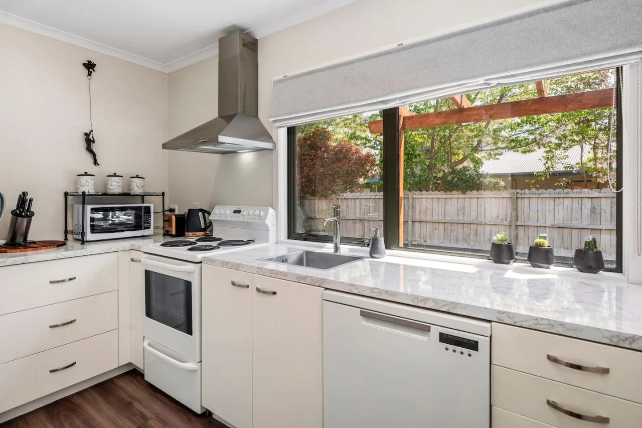 Kitchen with white cabinets, marble countertop, stove, oven, microwave, coffee maker, and a large window overlooking a backyard with a wooden fence and trees.