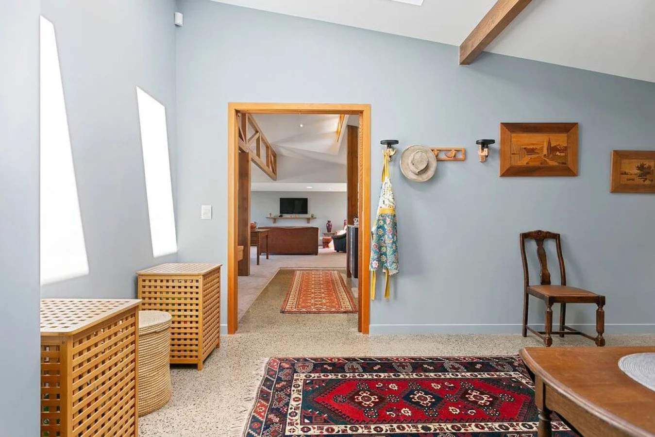 Interior view of a living space with light blue walls, wooden accents, and a patterned rug. A doorway leads to another room with a sofa and a wall-mounted TV. Decorative paintings and hats are hung on the wall, and a chair is positioned against the w