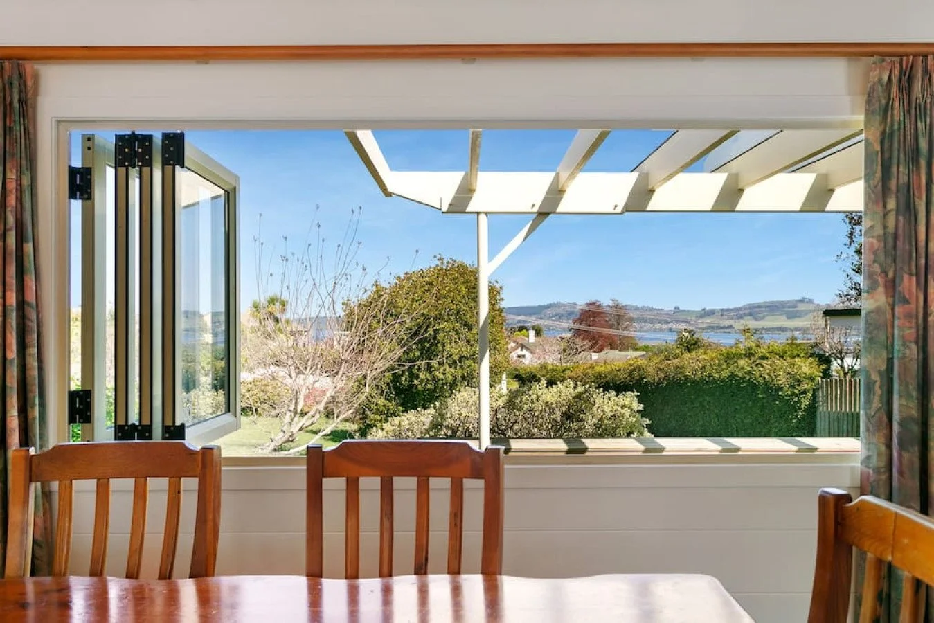 Interior view of a dining room with a large open window revealing a garden and scenic landscape with rolling hills and water in the distance.