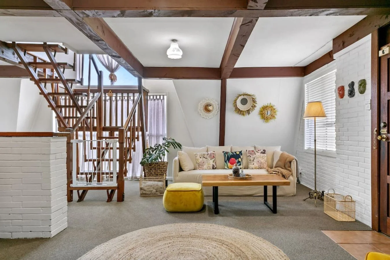 Living room with white sofa, decorative pillows, wooden coffee table, yellow ottoman, indoor plants, wall art, lamp, brick wall, staircase, window with blinds, and woven baskets.