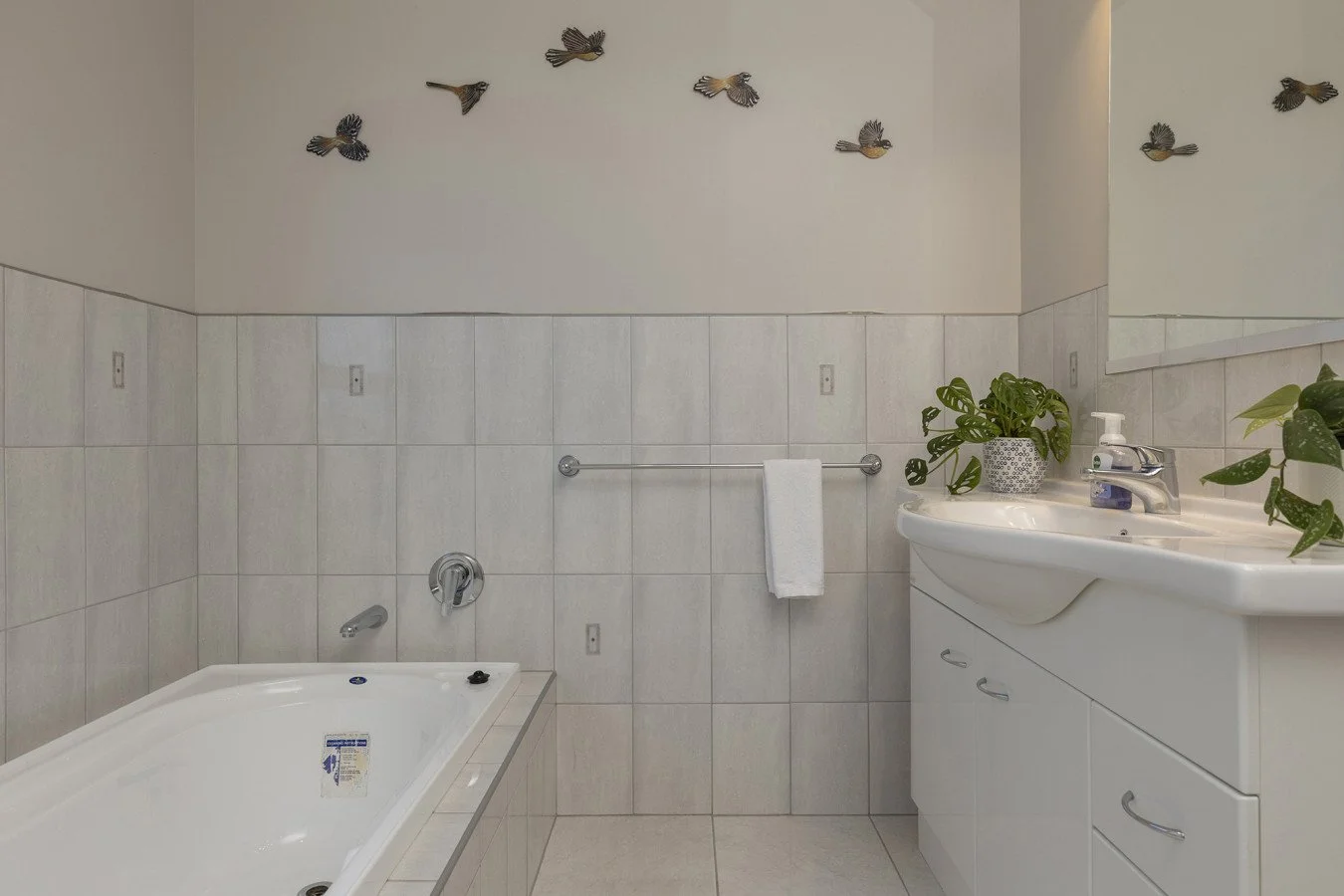 Bathroom with a corner bathtub, a white sink vanity with potted plants, a towel rack with a white towel, decorative bird wall art, and tiled walls.