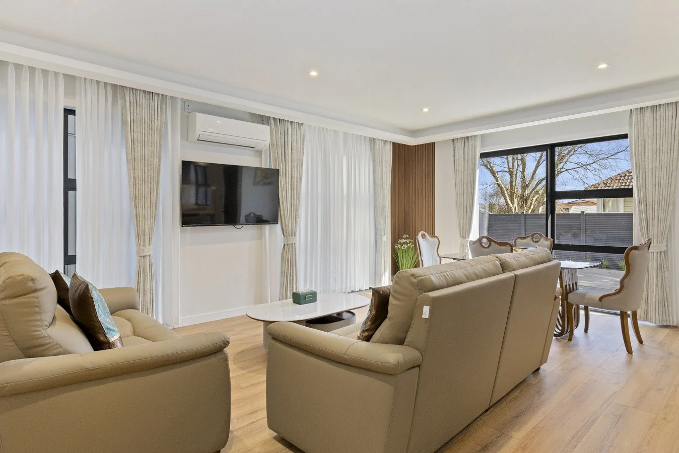 Bright living room with beige sofas, a wall-mounted flat-screen TV, large windows with white and beige curtains, a round dining table with white chairs, and wood flooring.