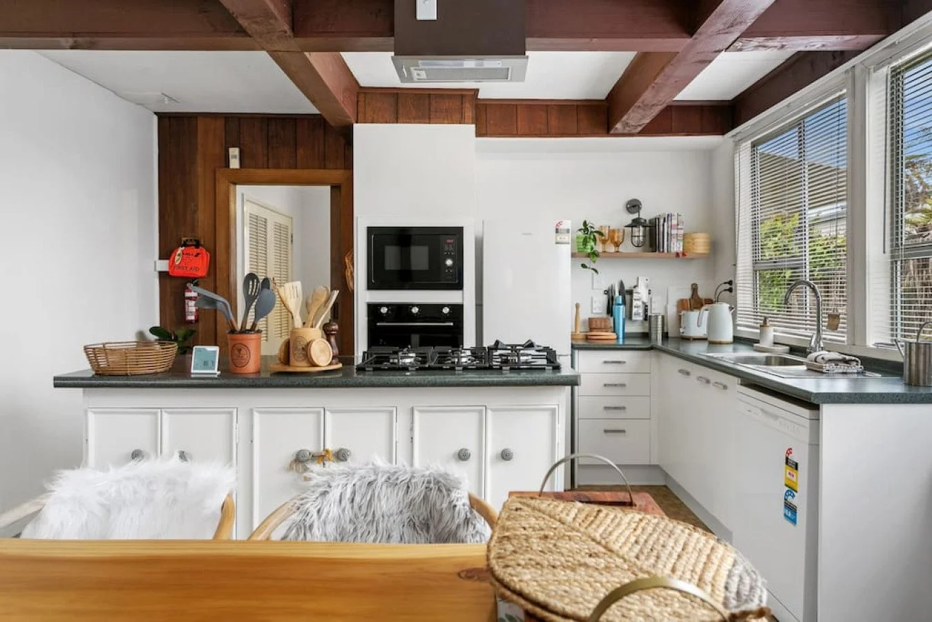 Kitchen with white cabinets, black countertops, a window with blinds, and various kitchen appliances and utensils, with part of a wooden table and chairs in the foreground.