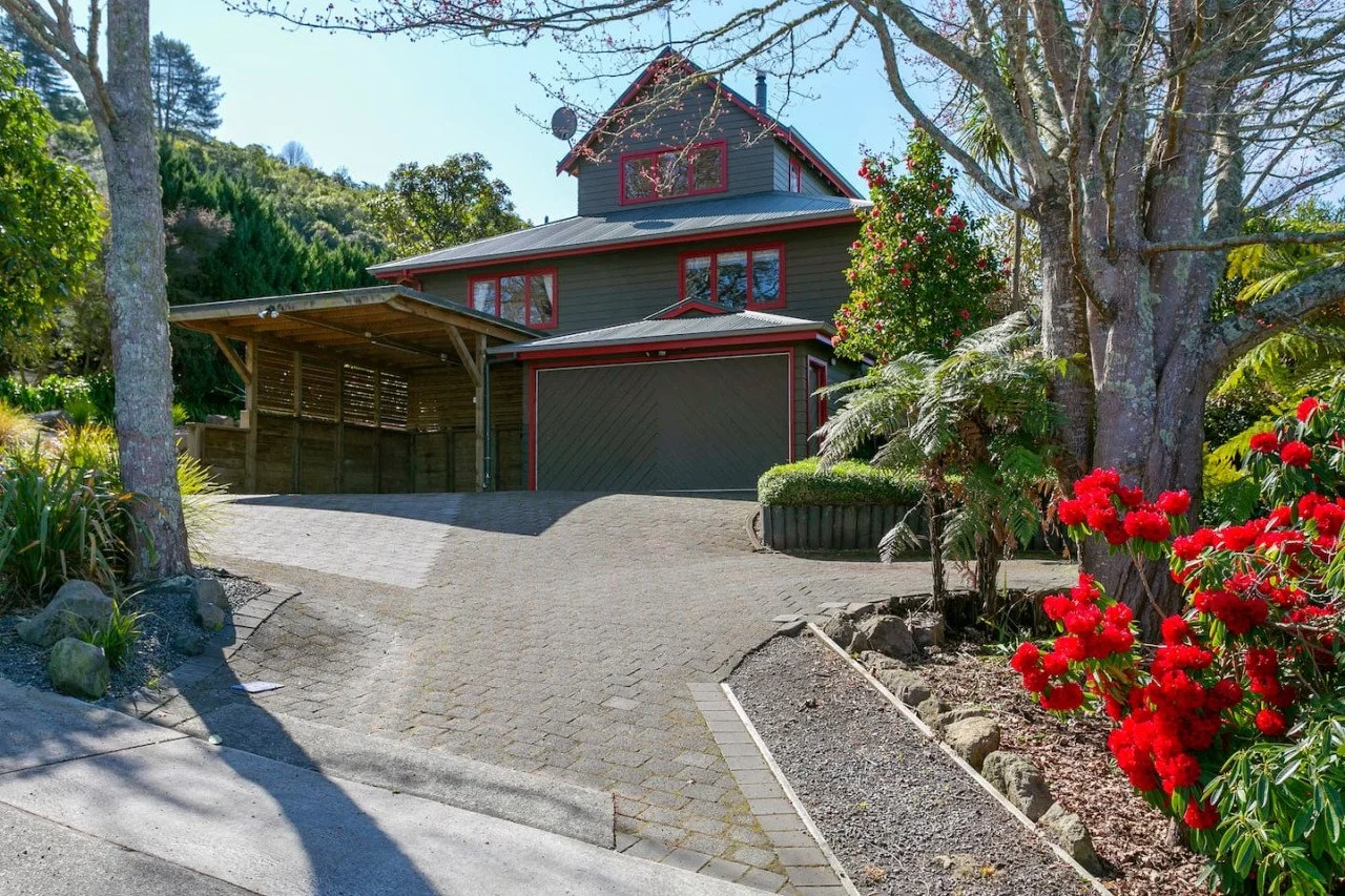 A house with dark gray siding and red window frames, a metal roof, and an attached carport, surrounded by trees and plants with red flowers, on a sloped paved driveway.