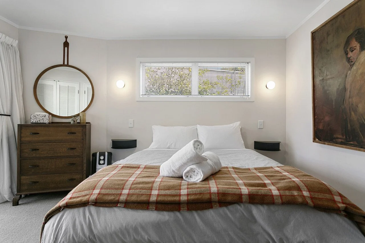 A neatly made bed with white pillows and a blanket. Towels are rolled and placed on top of the bed. There is a dresser with a mirror to the left, and a window above the bed. The room has artwork on the wall and soft lighting.