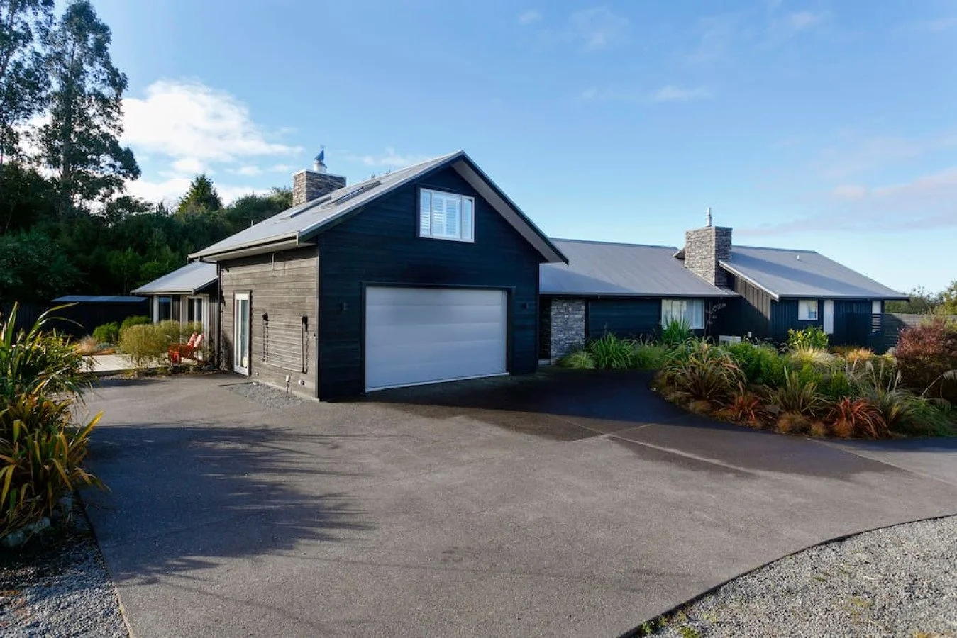 Modern black house with a white garage door, stone chimney, surrounded by landscaped garden with various plants, on a paved driveway under a partly cloudy sky.