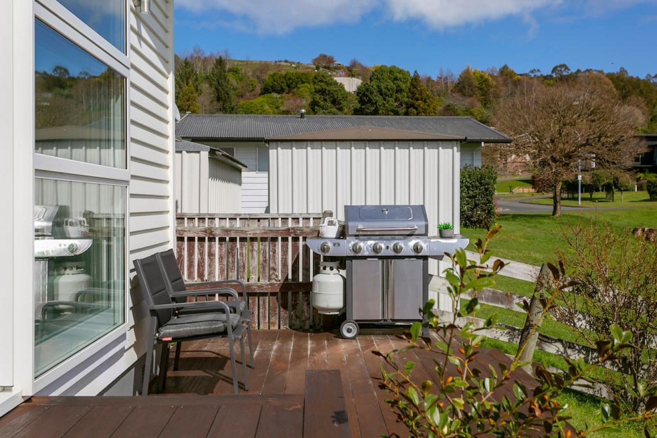 View of a backyard patio with a stainless steel barbecue grill, two black chairs, a small potted plant, and a grassy yard with trees and a road in the background.