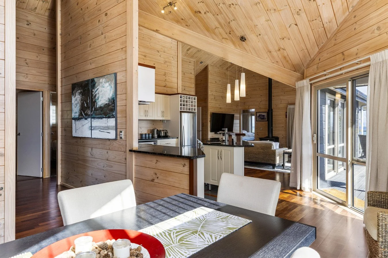 Wood-paneled living room with open kitchen, dining area, and large sliding glass door leading outdoor deck, furnished with white curtains and light-colored furniture.
