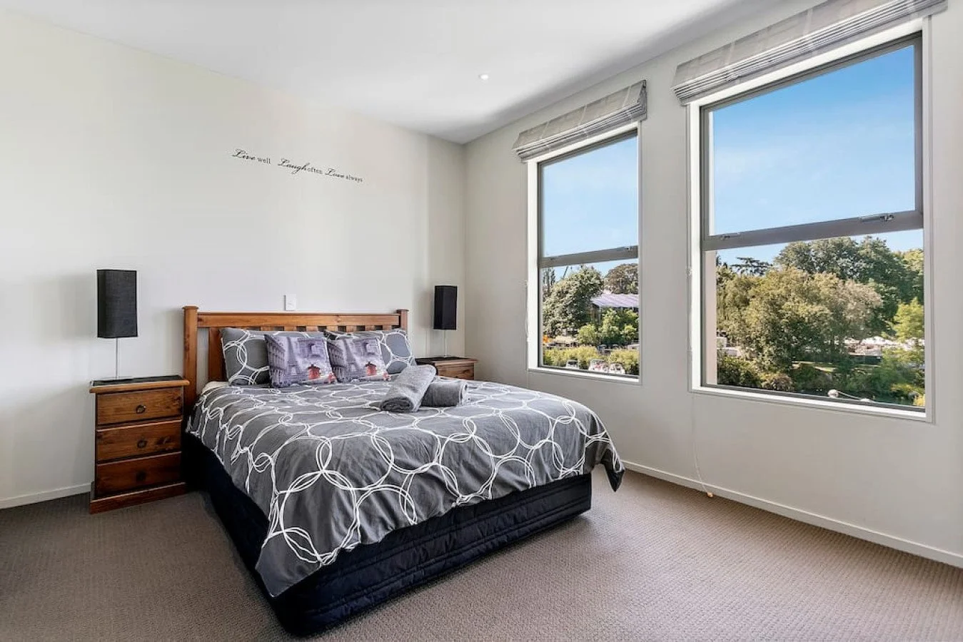 Bedroom with a bed, wooden nightstands, two black wall-mounted lamps, large windows showing trees outside, and a wall quote, 'Live Well Laugh Often Love Much'.