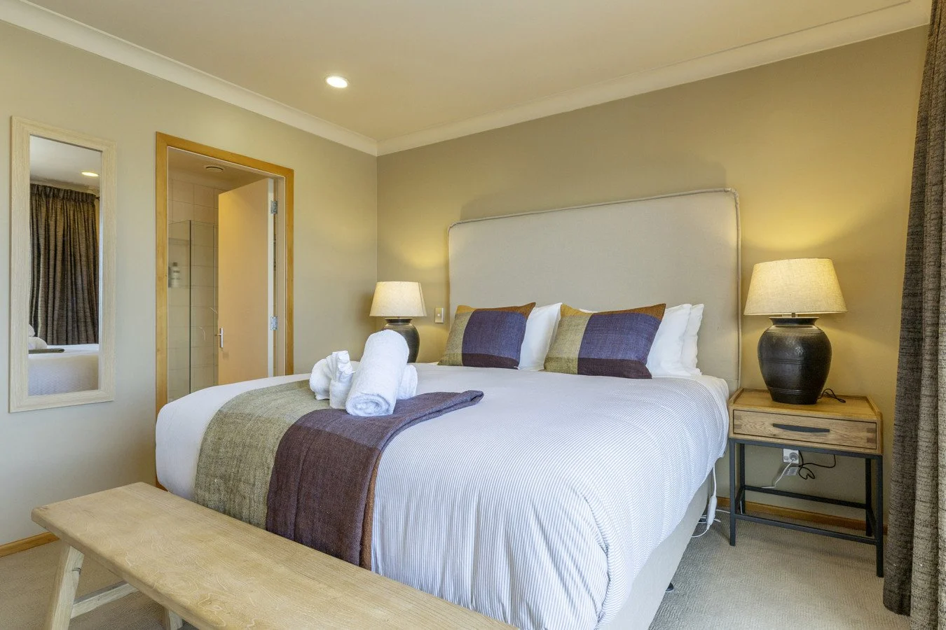 A neatly made hotel bedroom with a large bed, beige headboard, and decorative pillows, flanked by two bedside tables with lamps, and a towel arrangement on the bed.