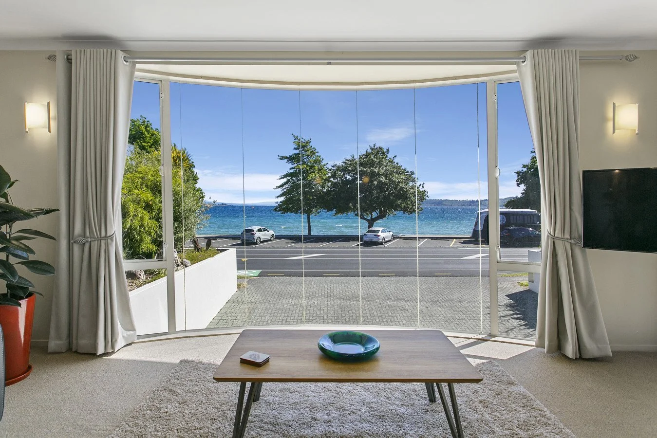 Living room with large glass window showing a view of the ocean, a parking lot, and trees outside. Interior includes white curtains, a wooden table with a green bowl and a book, a plant in an orange pot, and a wall-mounted TV.