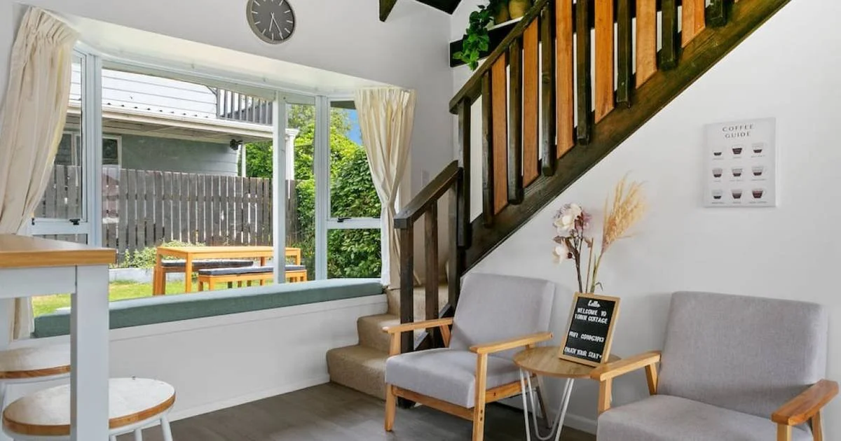 Living room with window seat, two gray armchairs, wooden side table with a blackboard sign, staircase, and wall art.