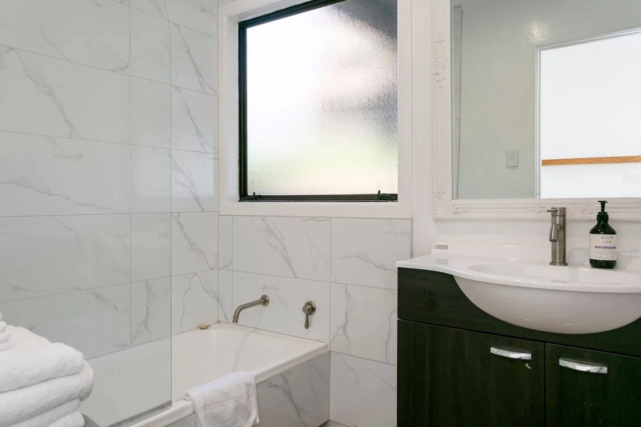 Bathroom with a white bathtub, frosted window, dark wood vanity with a white sink, and a mirror, with rolled white towels on the bathtub