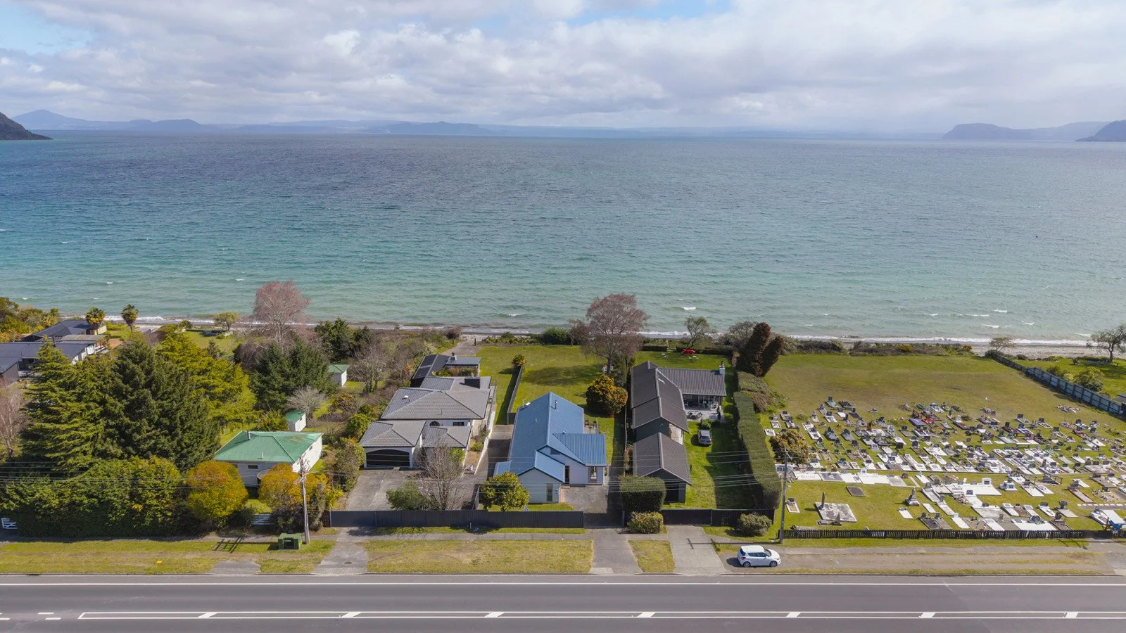 An aerial view of residential homes and a cemetery by the beach with mountains in the background.