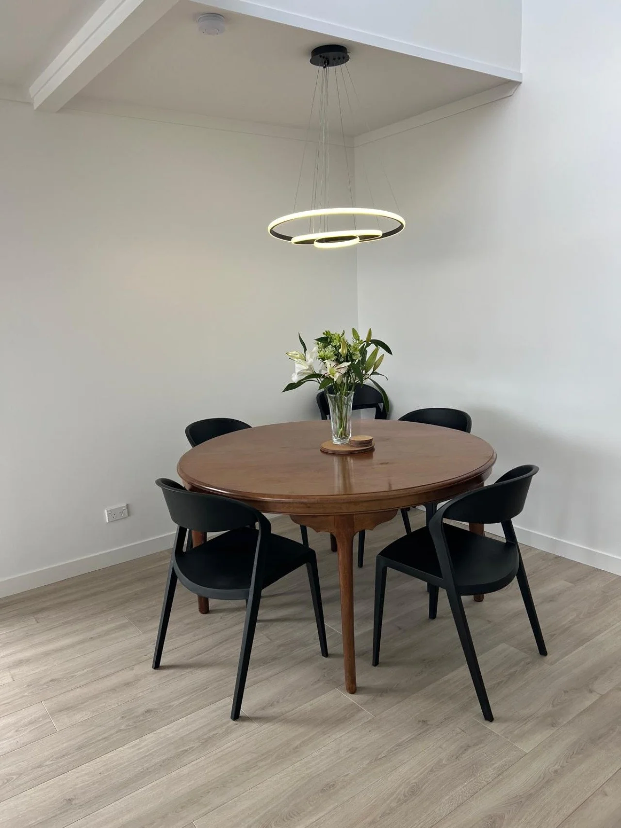 Dining room with a round wooden table, four black chairs, a vase with white lilies, a modern circular pendant light fixture, and light wood flooring.