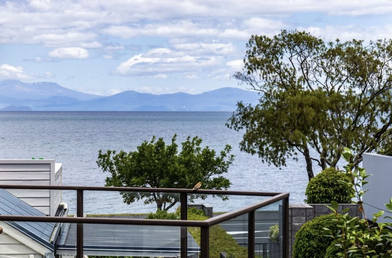 View of a lake with mountains in the background, framed by patio railing, trees, and shrubs in the foreground.