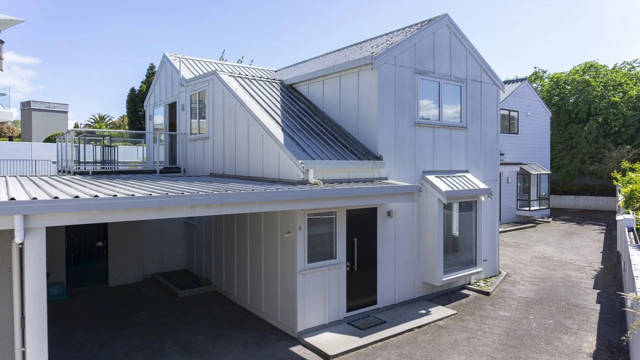 A white multi-story house with metal roofing, multiple windows, and a small balcony on the upper floor, situated in a sunny area with a paved driveway and green trees in the background.