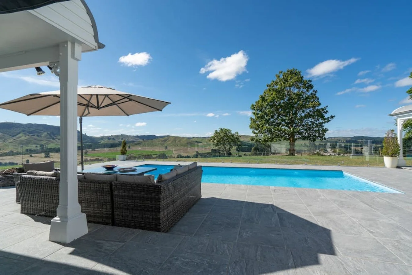 Poolside patio with outdoor seating, umbrella, and views of rolling green hills and trees under a blue sky with clouds.