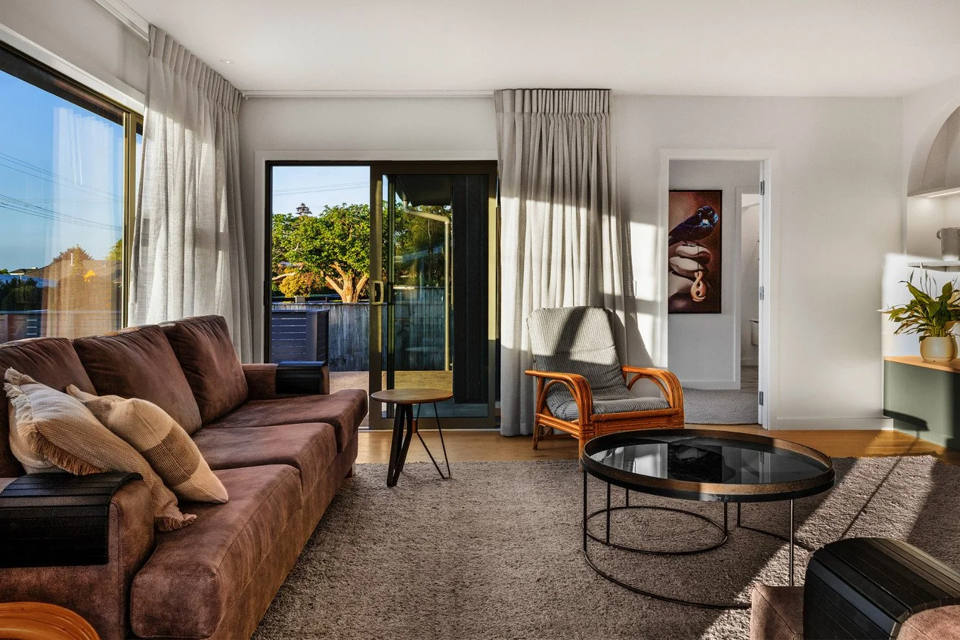Living room with a brown couch, a small side table, a gray armchair, a black round coffee table, and a view through a sliding glass door to a backyard with trees.