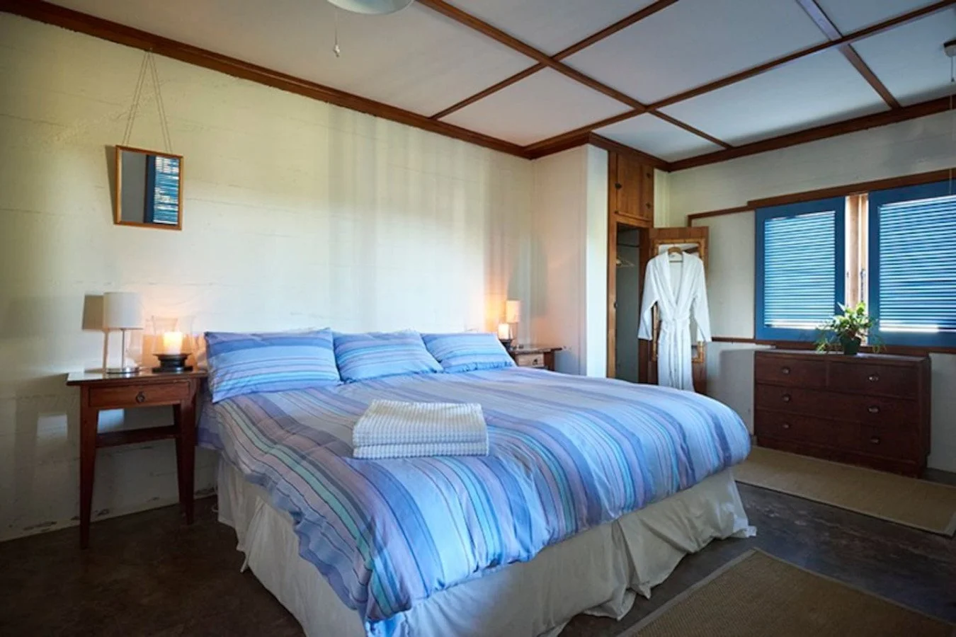 A cozy bedroom with a large bed covered in a blue and purple striped comforter, two bedside tables with lamps, a wooden dresser near the window, a white bathrobe hanging on a closet door, and a ceiling with wooden accents.
