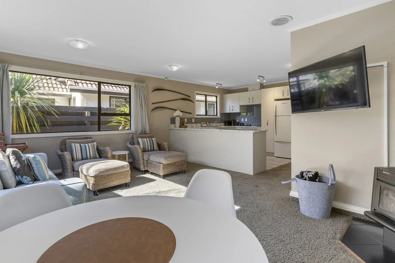 Living room and kitchen with beige walls, gray carpet, and large window with curtains. Furnished with chairs, sofa, TV on wall, and kitchen appliances.