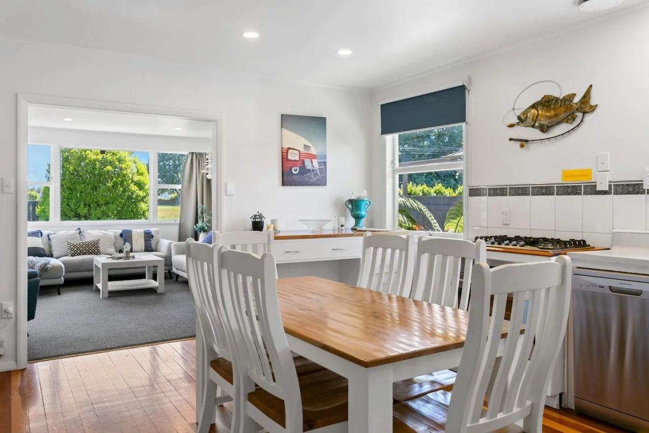 Kitchen and living room with white walls, wooden dining table with six white chairs, kitchen counter with a stove, decorative fish mounted on the wall, and a living room with white sofas and large windows showing green trees outside.