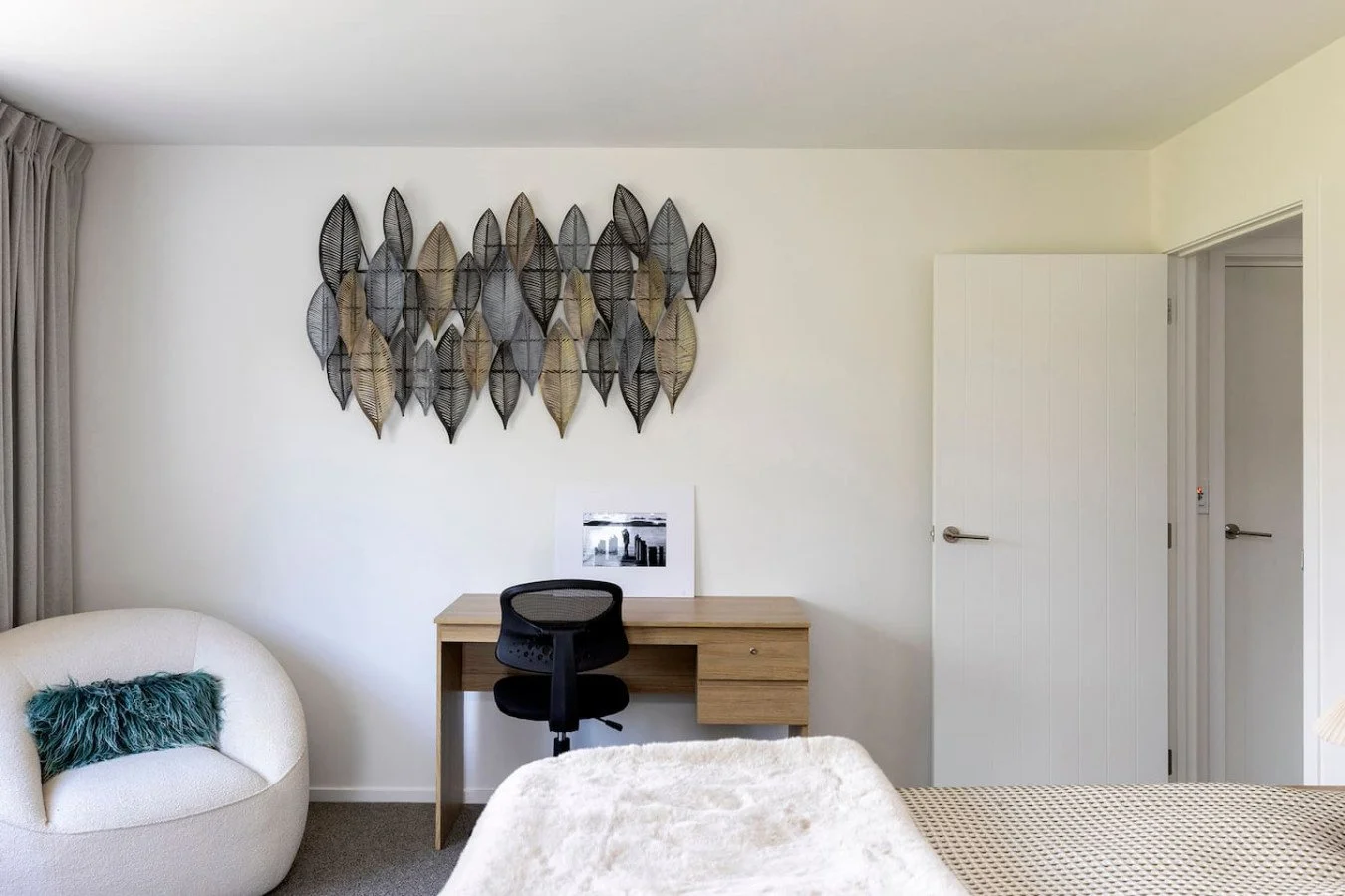 Bedroom with white walls, a black desk with a chair, a frame with black and white art, a curved white armchair with a green pillow, and decorative wall art made of overlapping metallic leaves.