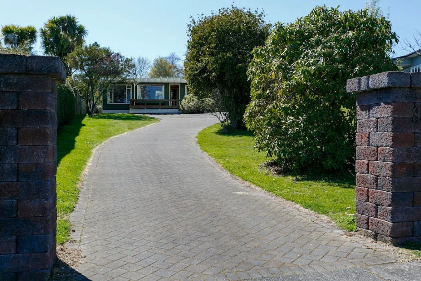 A curved driveway leading to a house, flanked by green grass and shrubbery, with brick pillars at the entrance on a sunny day.