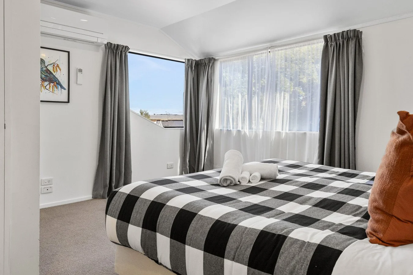 A neatly made bed with a checkered black and white bedspread, orange pillows, and folded white towels on top, in front of large windows with gray curtains in a bright, modern bedroom.