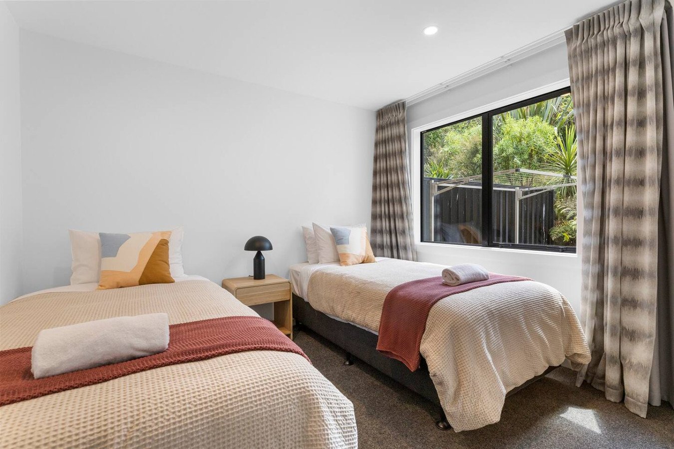 A hotel room with two single beds, beige bedspreads, and pillows, two rolled towels on each bed, a nightstand with a black lamp, a large window with beige curtains, and greenery outside.