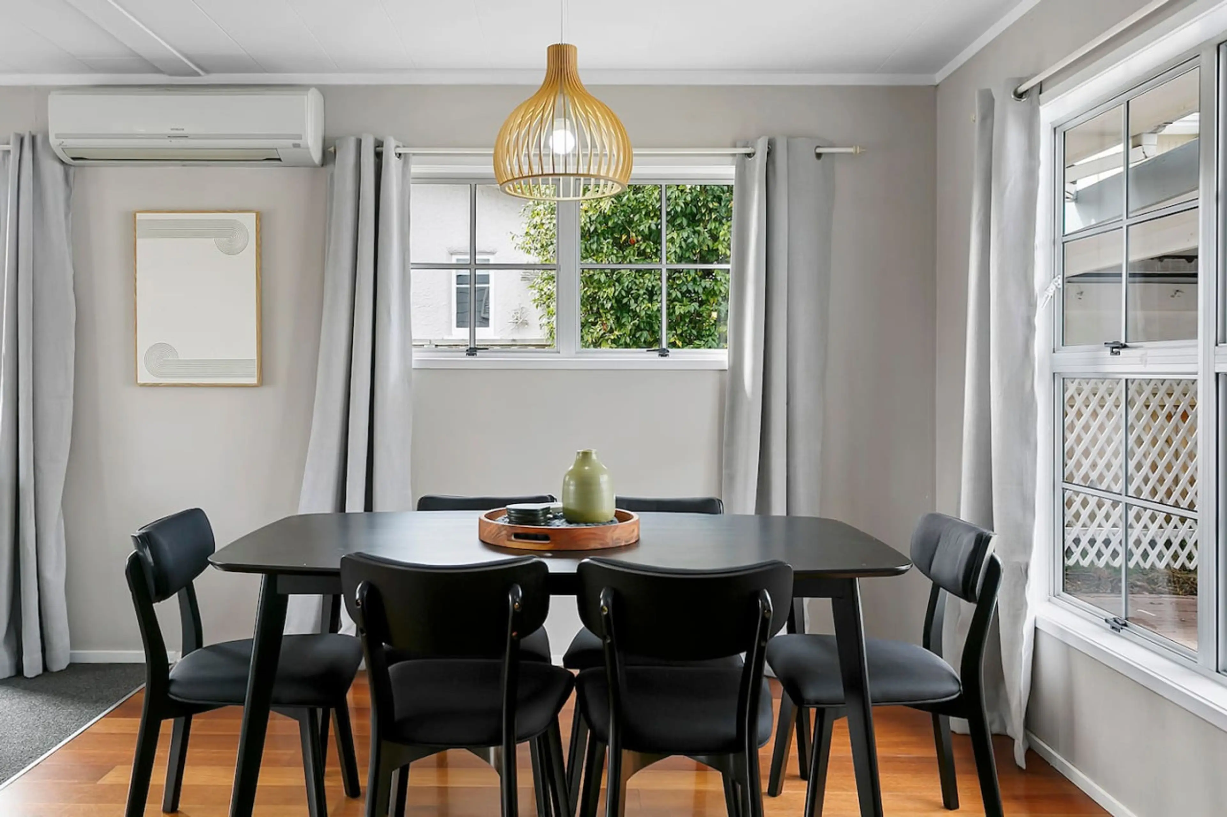 Dining room with black table and chairs, white curtains, window showing outdoor greenery, wooden pendant light, wall-mounted air conditioner, framed artwork on wall, wooden floor.