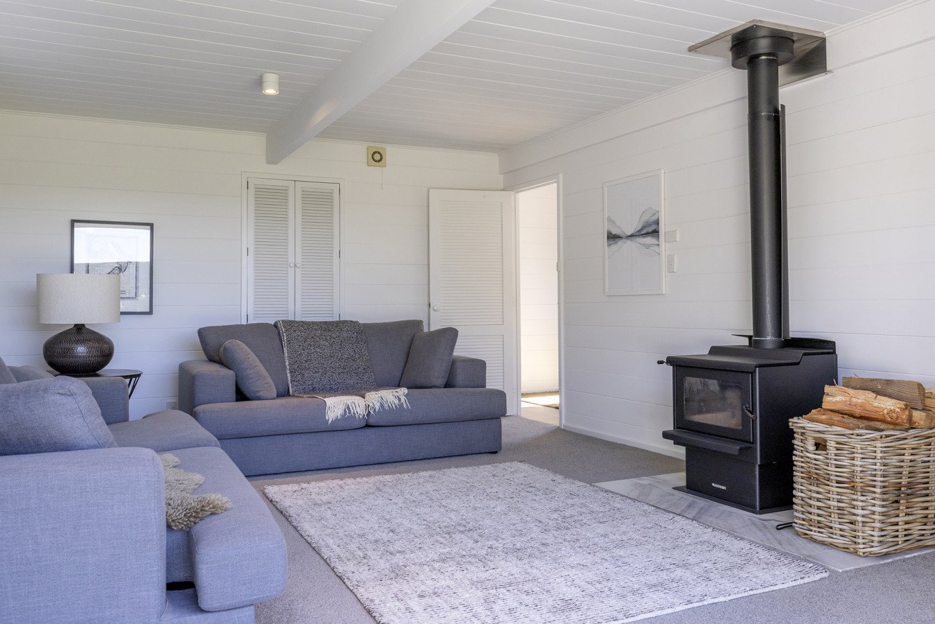 Living room with gray sofas, a black wood stove, a table lamp, and a framed picture on white paneled walls.