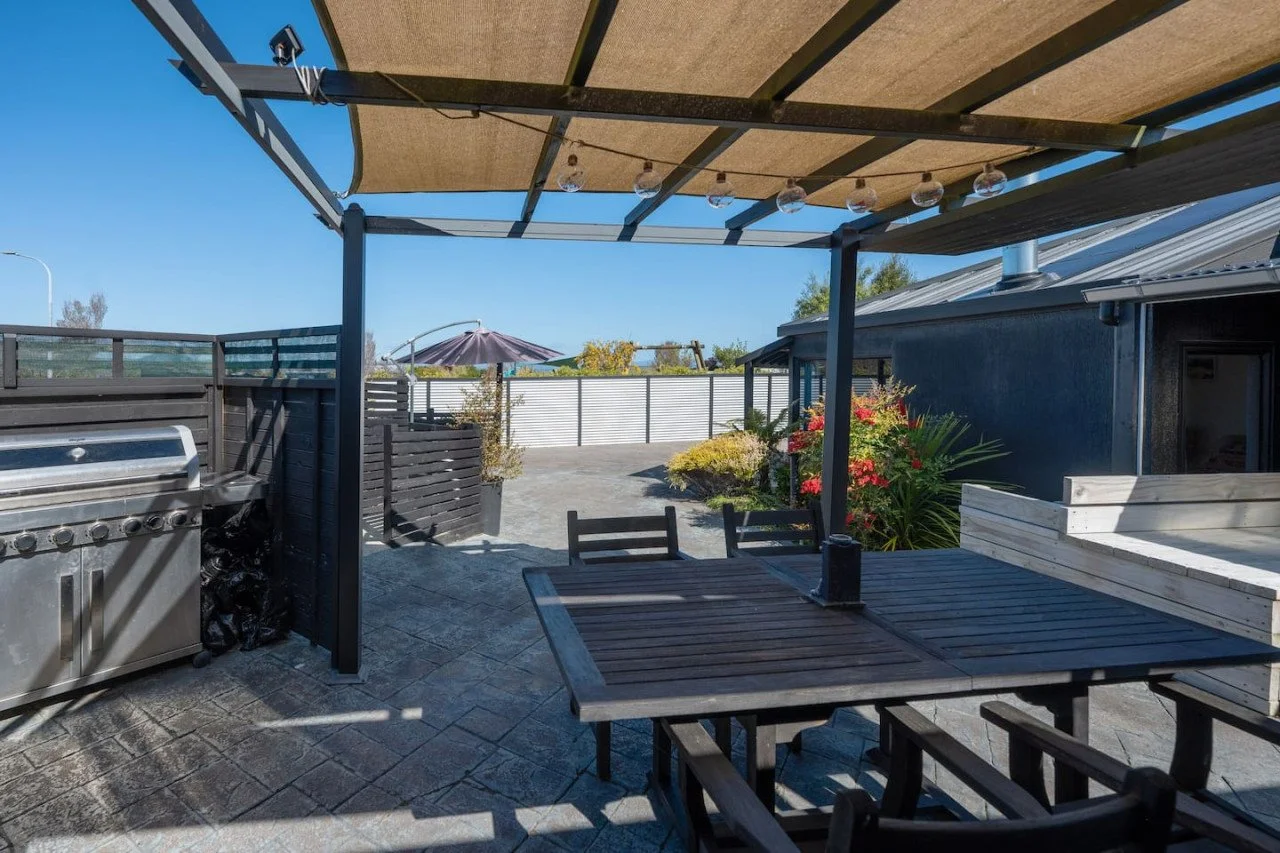 Outdoor patio area with a barbecue grill, wooden dining table with chairs, potted plants, flower bushes, an umbrella, and a fenced yard under a blue sky.