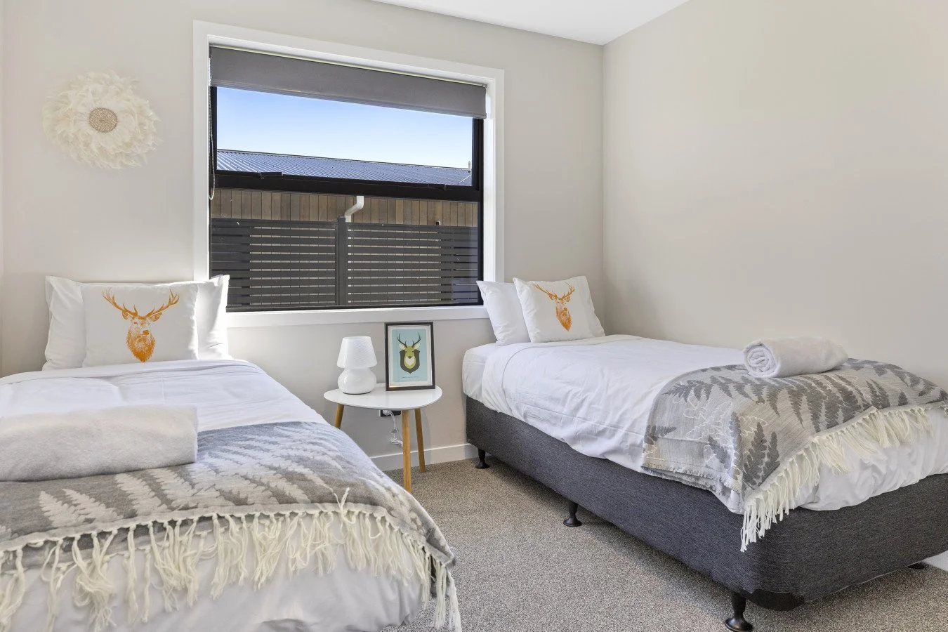 A bedroom with two beds, white bedding, decorative pillows with deer head designs, a window with a view outside, a small white side table with a lamp and framed deer art, and a neutral-colored carpet.