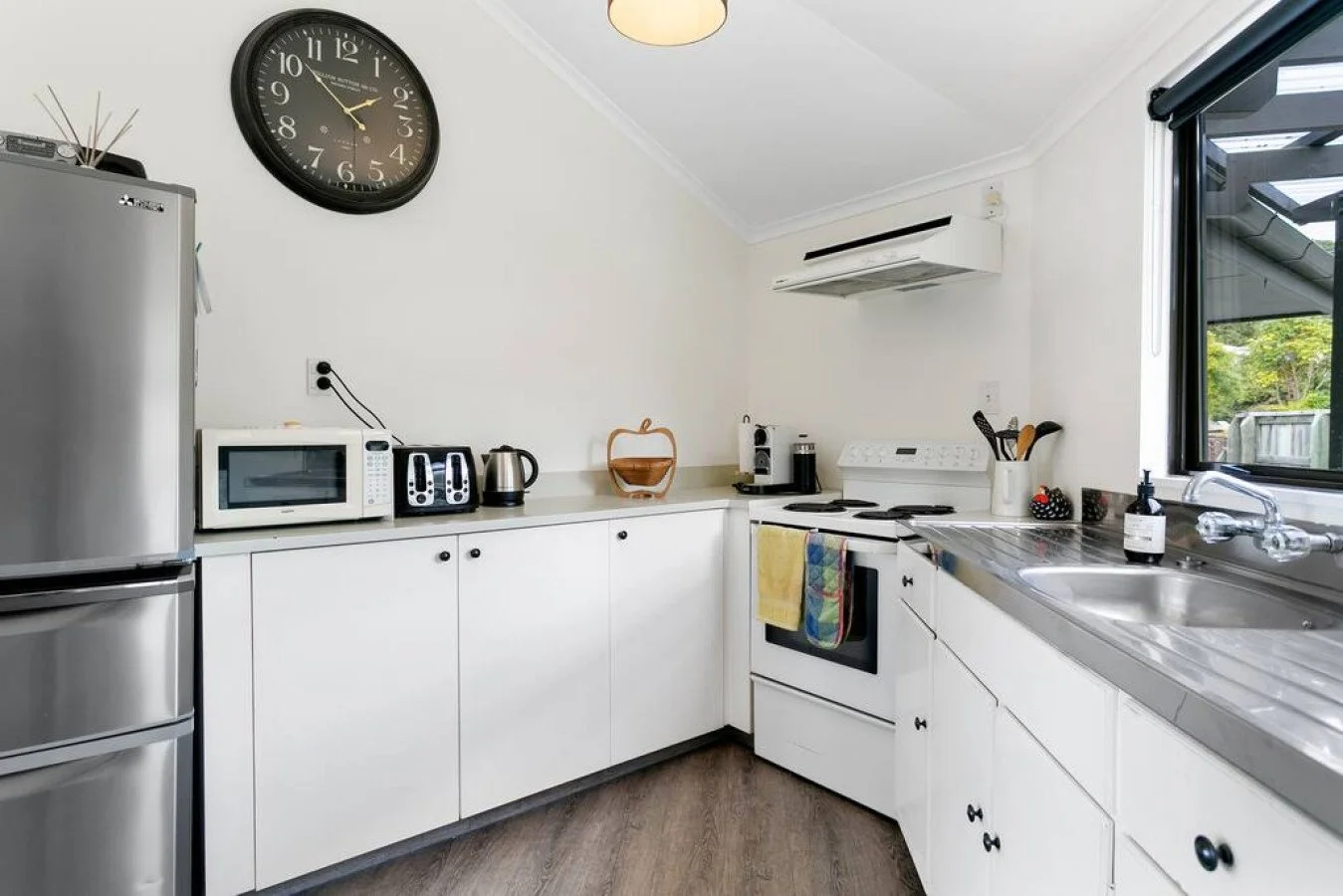 A kitchen with white cabinets, a stainless steel fridge, microwave, toaster, electric kettle, and coffee machine on the counter. A large wall clock, air conditioner, and a window with a view of a backyard are visible.