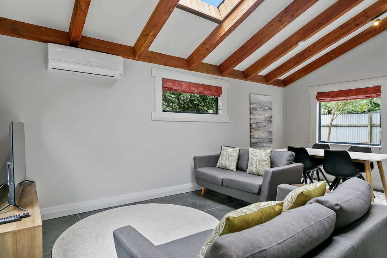 Living room with gray sofa, black dining chairs, wood ceiling beams, windows with red valances, white walls, and hardwood floor.