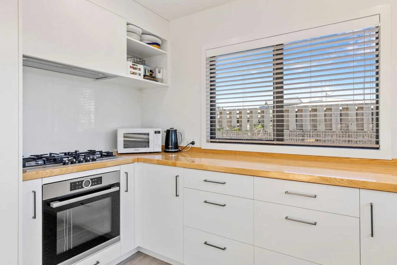 White kitchen with wooden countertop, gas stove, oven, microwave, black electric kettle, and window with blinds.