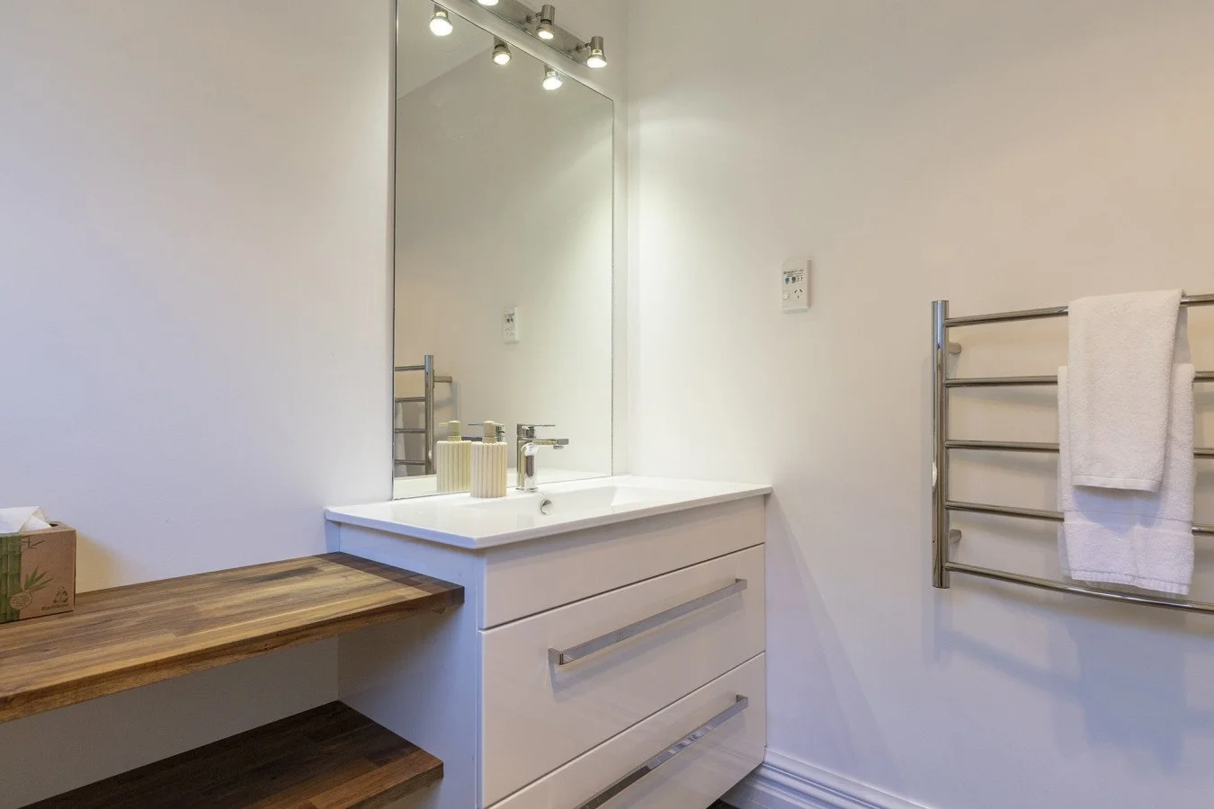 Modern bathroom with white walls, a large mirror, a white vanity with a sink and three drawers, a wooden countertop, two soap dispensers, an electric switch or thermostat on the wall, and a stainless steel towel heated rack with a white towel.