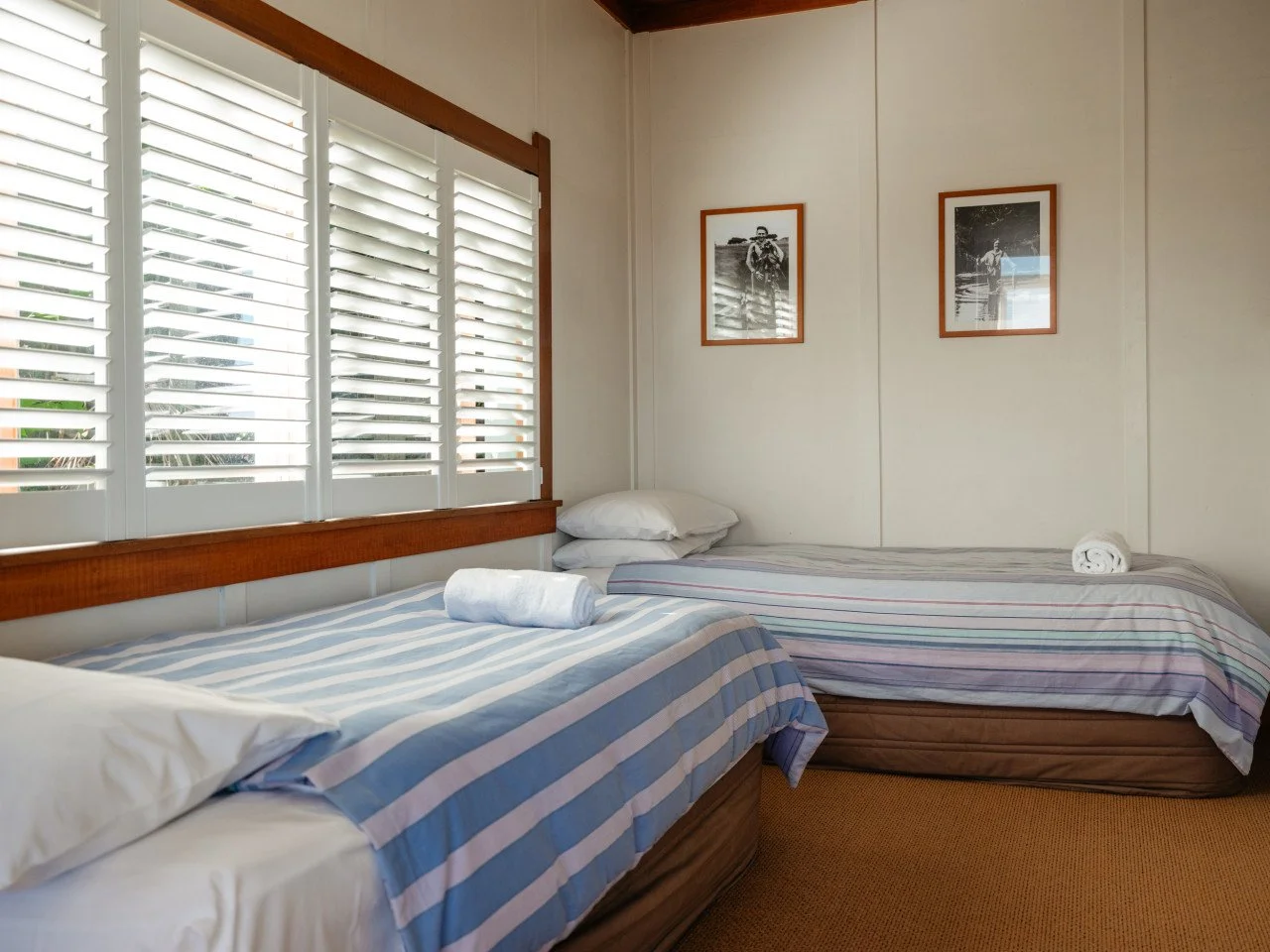 A bedroom with two single beds with striped bedding, pillows, and rolled towels, near a large window with white plantation shutters, and two framed black-and-white photos on the wall.
