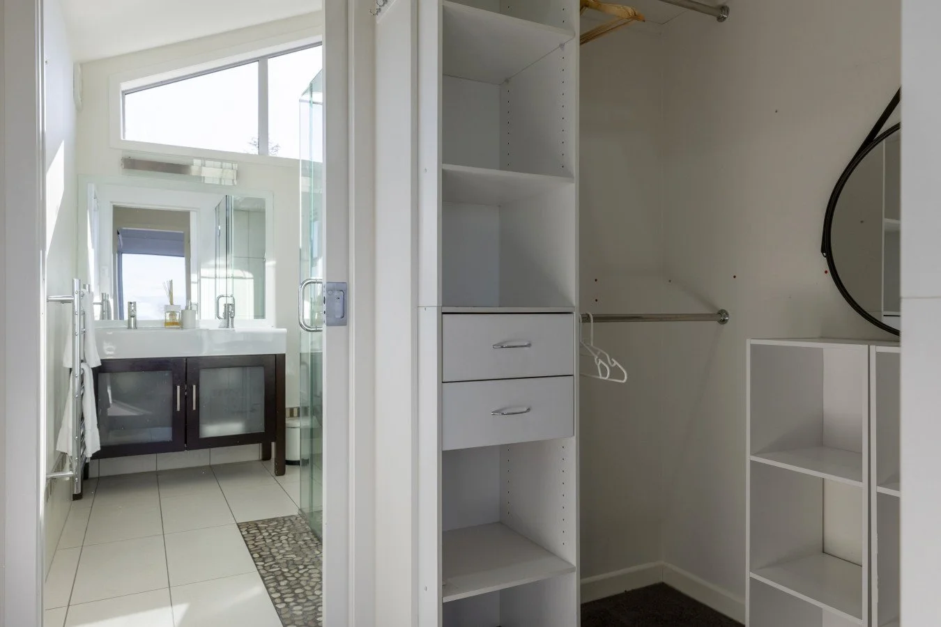 Empty walk-in closet with built-in shelves, drawers, and hanging rod, adjacent to a bathroom with a vanity, mirror, and window.