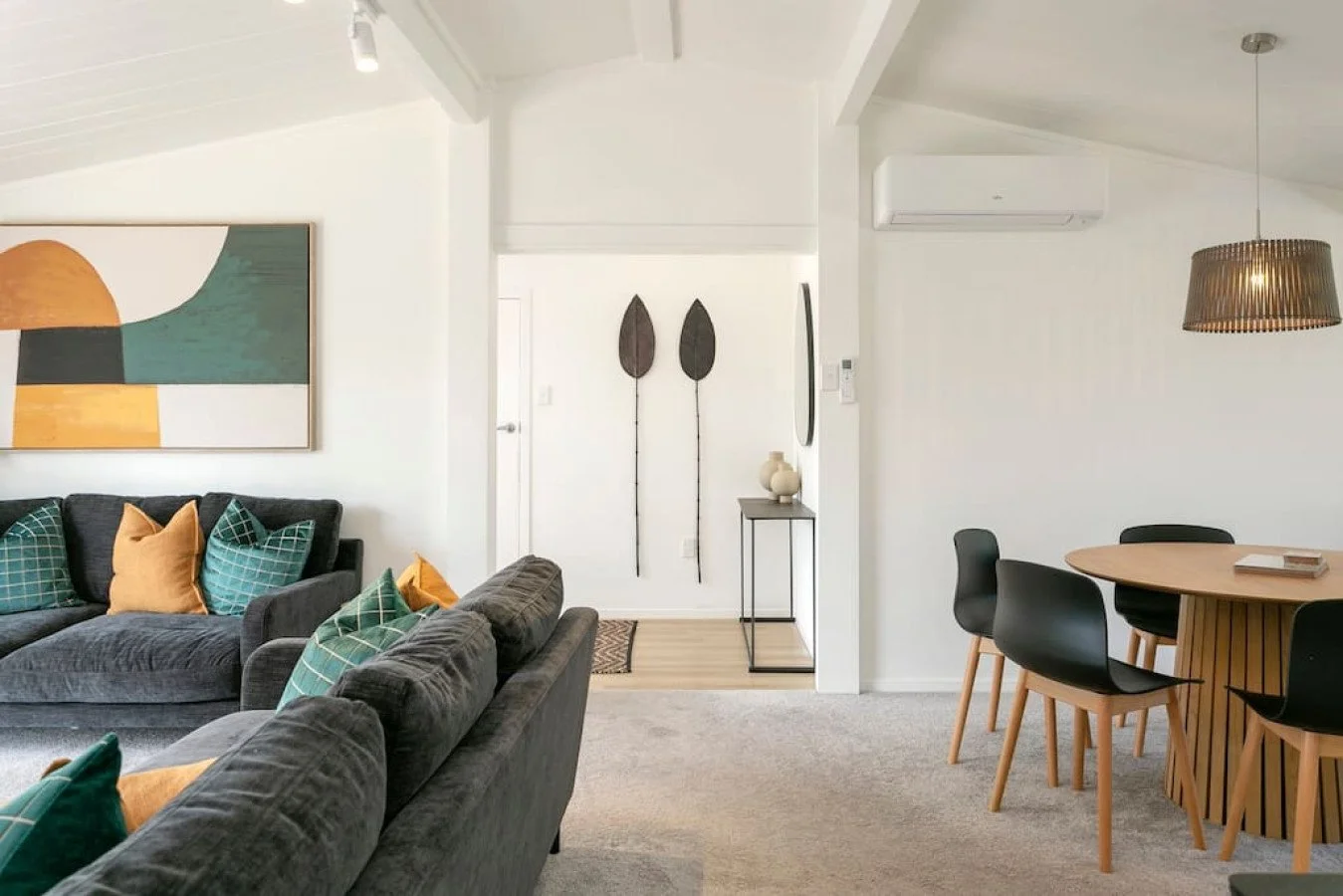 Living room with dark gray sofas, decorative pillows, abstract wall art, and a wooden dining table with black chairs, light-colored walls, and pendant light.