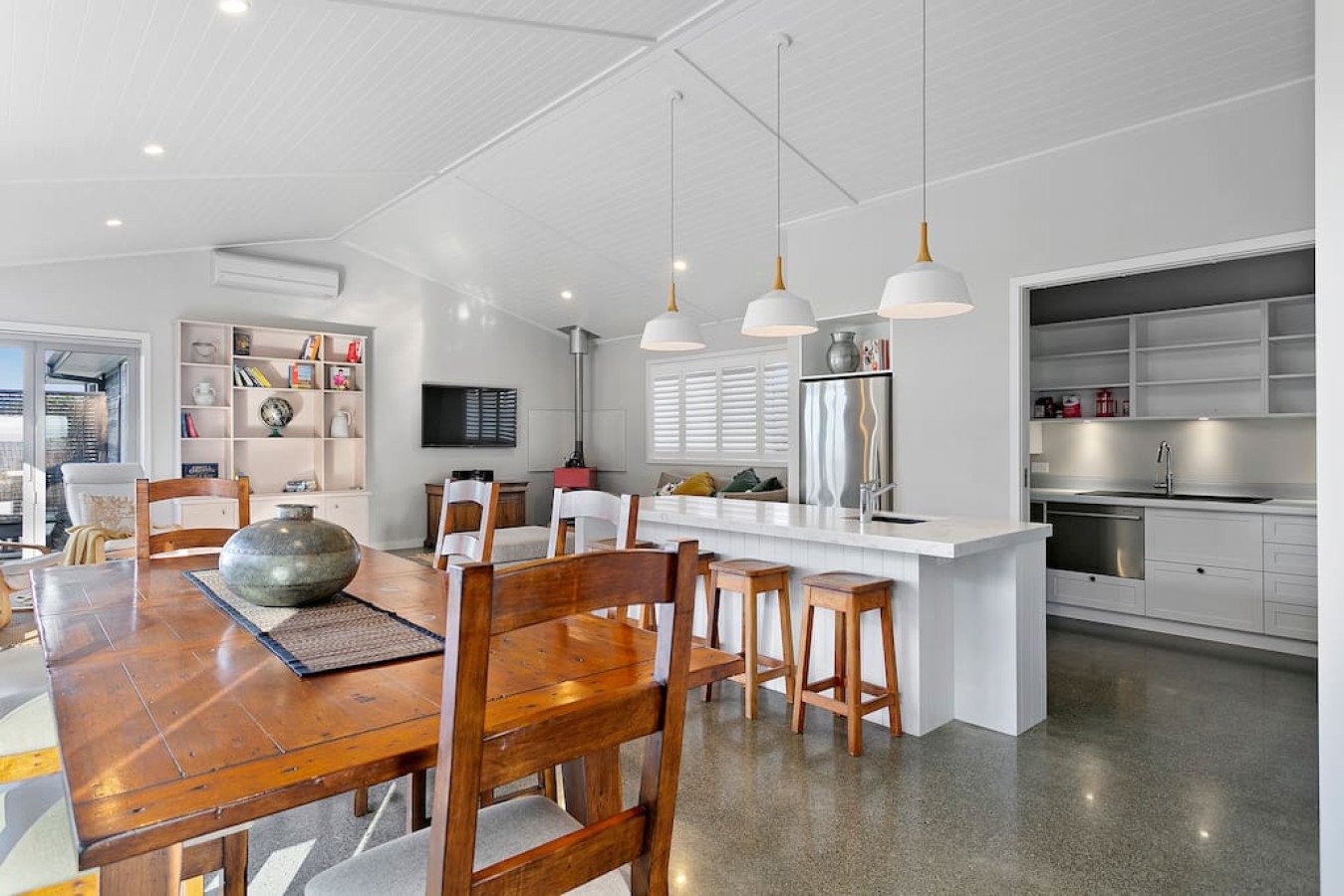 Living and dining area with a wooden dining table, white walls, a kitchen bar with stools, a couch, a wall-mounted TV, white shelves, and a kitchen with white cabinets and stainless steel appliances.