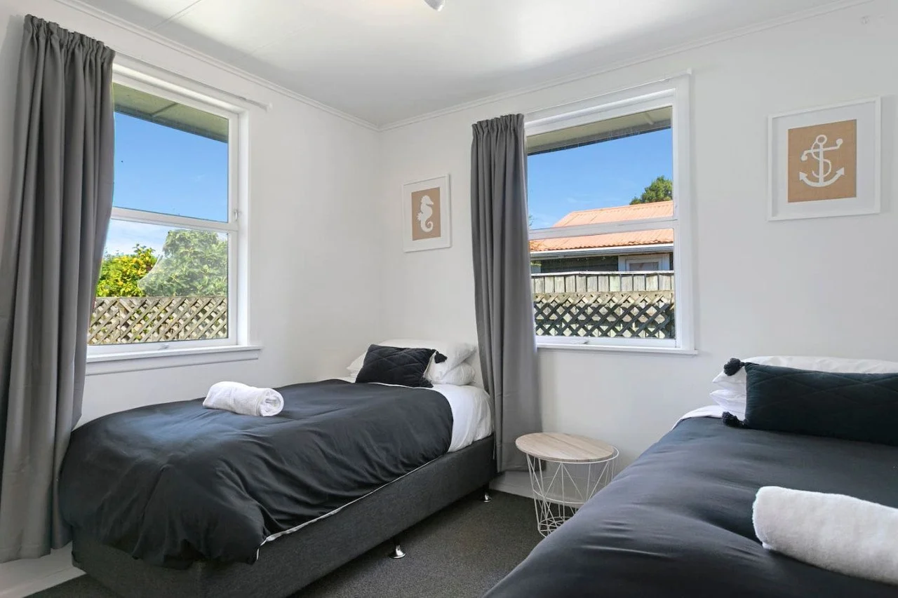 Two twin beds in a bright, modern bedroom with white walls and gray bedding, each with rolled towels and black pillows, near windows with gray curtains, outdoor view of trees and neighboring houses, framed nautical-themed artwork on the walls.