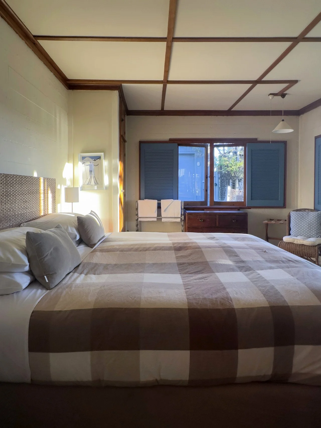 A cozy bedroom with a large bed covered in a plaid blanket, pillows, and a headboard, with wooden ceiling beams, double windows with blue shutters, a wicker chair, and a small dresser.