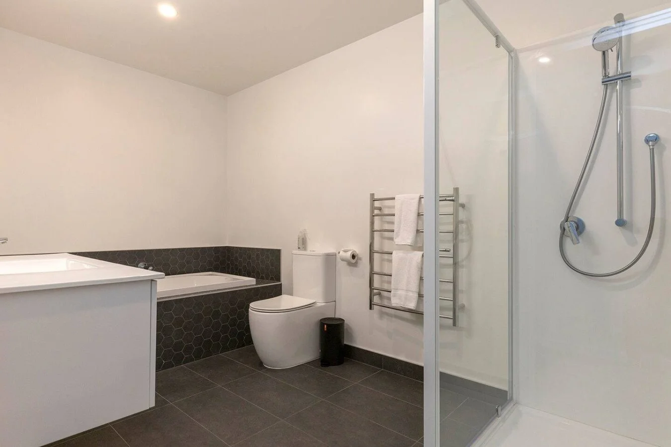 Modern bathroom with white walls, a bathtub with black hexagonal tiles, a toilet, a towel rack with towels, a shower stall with glass door, and dark tiled flooring.