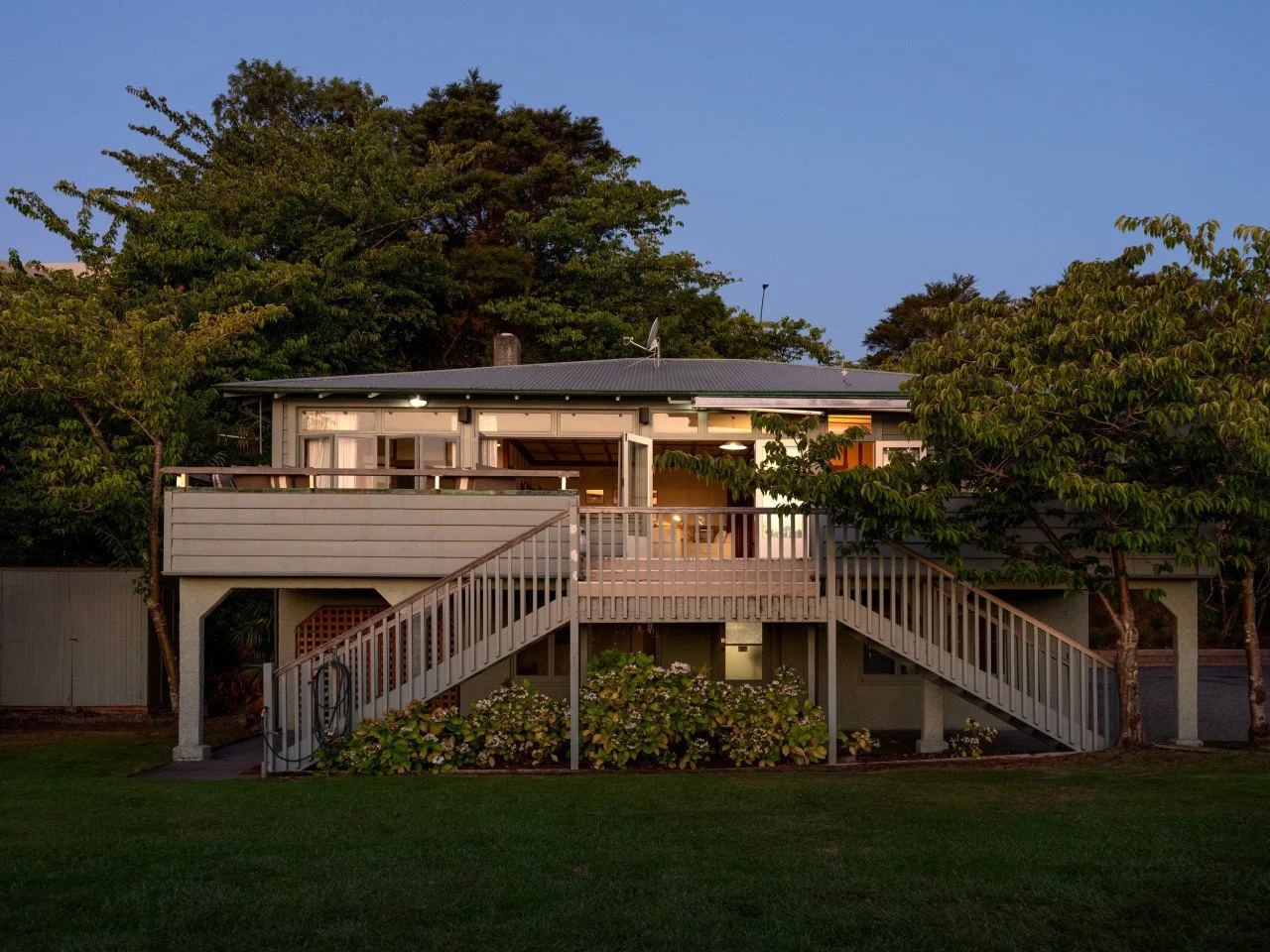 A house with a large deck, stairs on both sides, surrounded by trees and greenery during dusk.