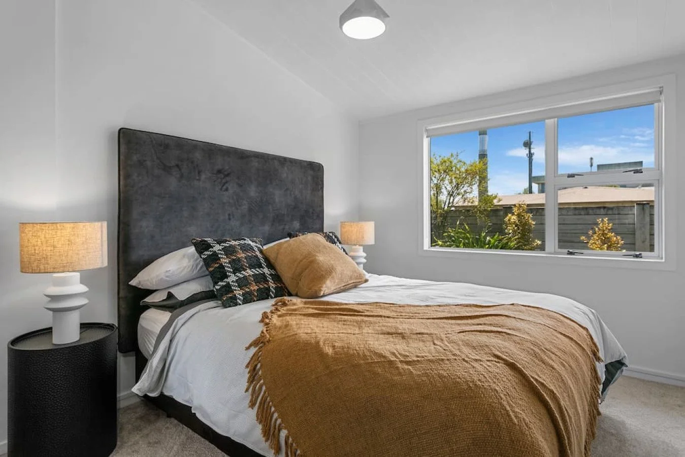 A bedroom with a large window showing trees and blue sky outside. The bed has a black headboard, white sheets, pillows, and a brown blanket. There are two bedside lamps on white nightstands.