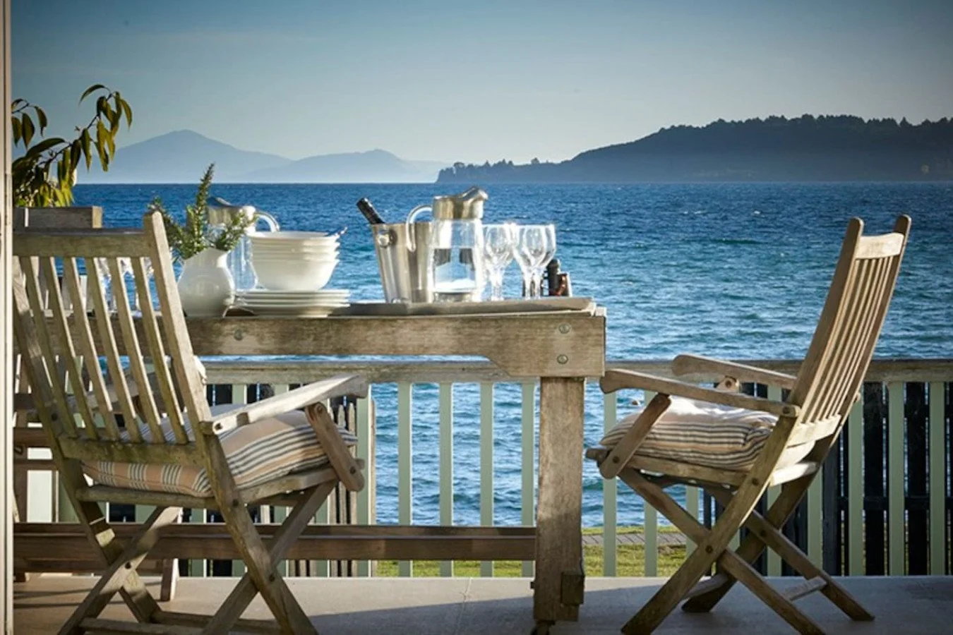 A wooden patio table set with wine glasses, bowls, and a wine cooler, overlooking a calm blue lake with distant mountains and a tree-lined shoreline in the background.