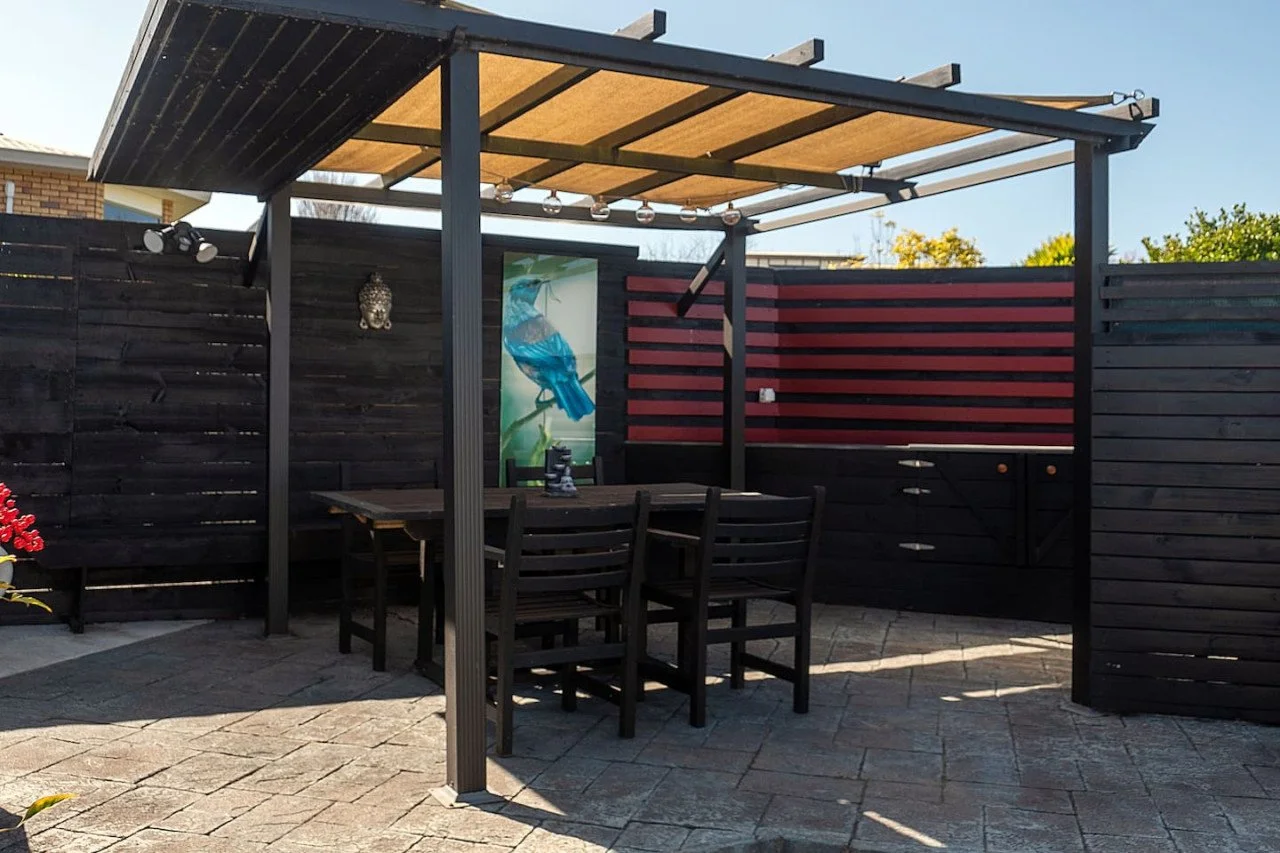 Outdoor patio area with a black wooden pergola, black dining table, and four matching chairs. The patio is enclosed by black and red slatted wooden fencing, with glass string lights hanging from the pergola and a decorative owl wall hanging on the bl