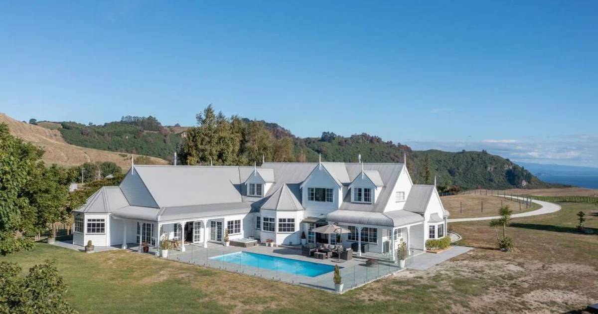 A large white house with multiple gables and a swimming pool in the backyard, surrounded by hills and open land under a clear blue sky.